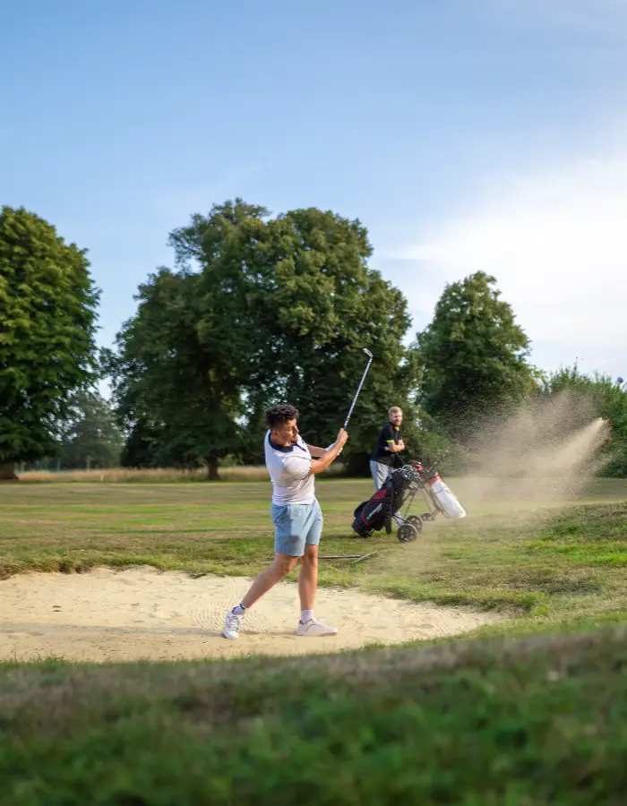 A young man, wearing a white t-shirt and blue short, is mid swing taking his shot from the golf bunker. A man in black watches from the side lines with two golf caddys.