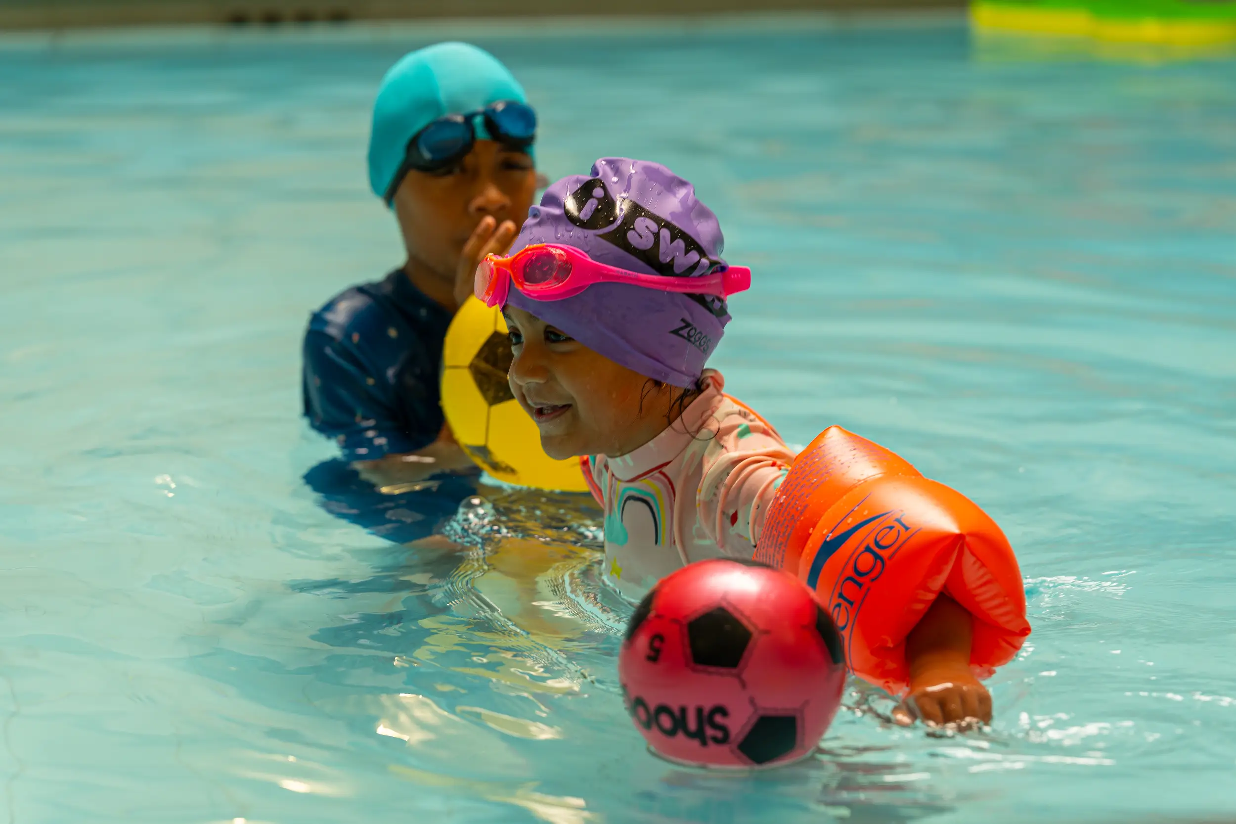 Two young children playing in a swimming pool, wearing colourful swim caps, goggles and armbands, smiling and holding foam balls.