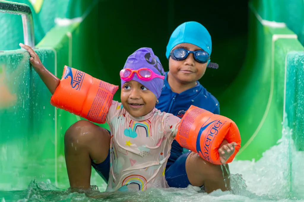 Two young children coming down a green water slide at an indoor swimming pool, wearing goggles and orange armbands.