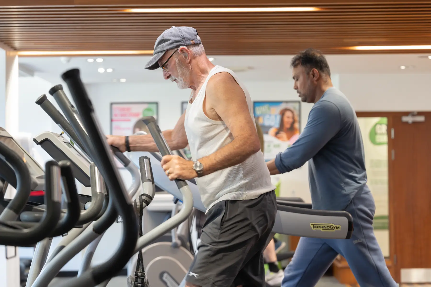 An older man, with a grey gap, using a cross trainer in the gym.