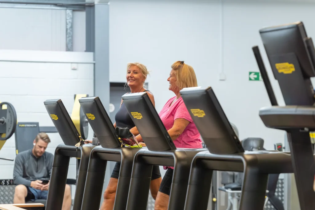 Two women smiling and chatting while using treadmills in a gym.
