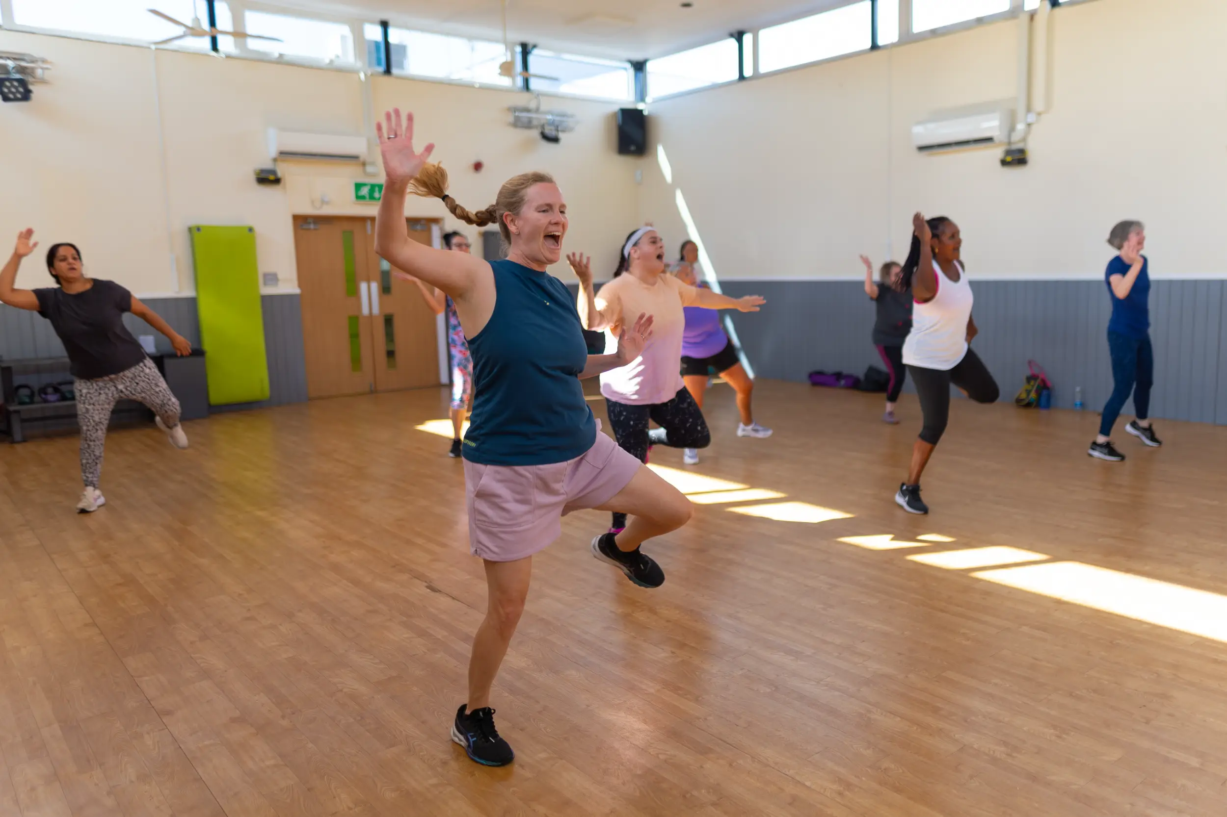 A group of women energetically taking part in a dance fitness class in a sports hall.