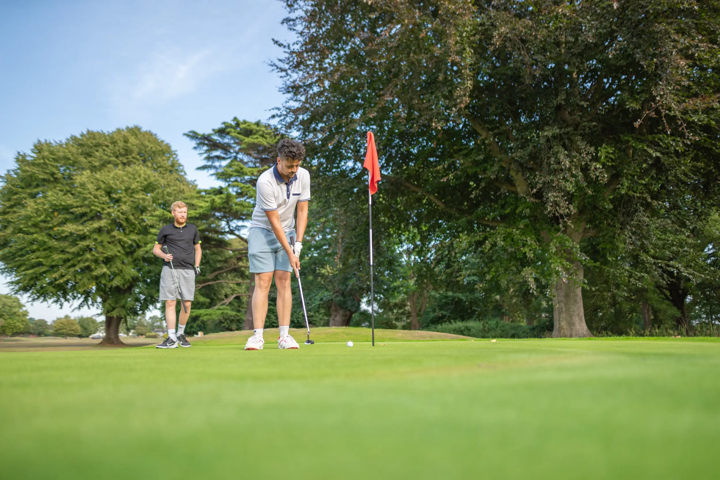 Two men playing golf on a green course surrounded by trees on a sunny day, one preparing to putt near the flag.