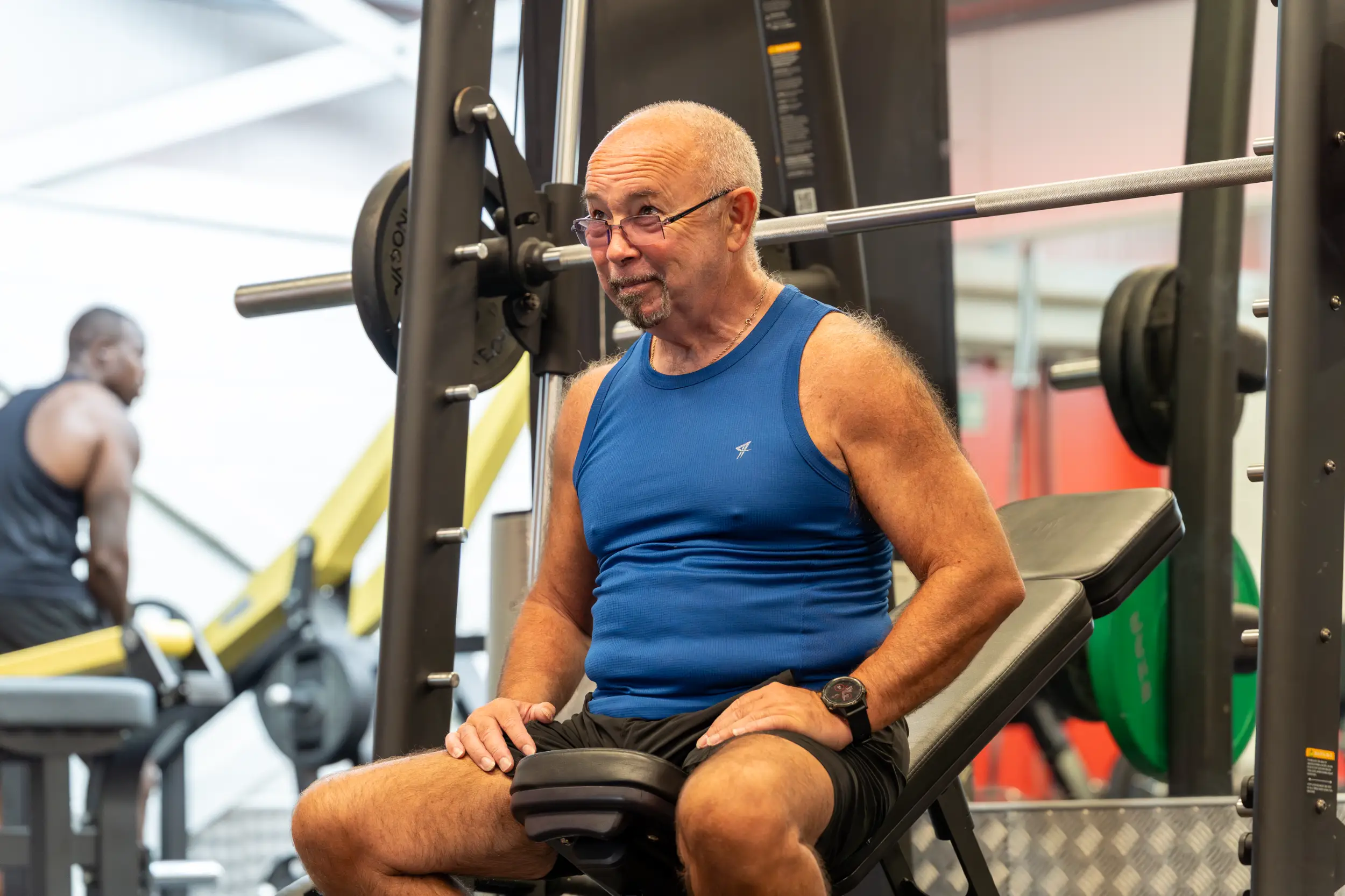 An older man in a blue vest top sitting at a weight machine in a gym.
