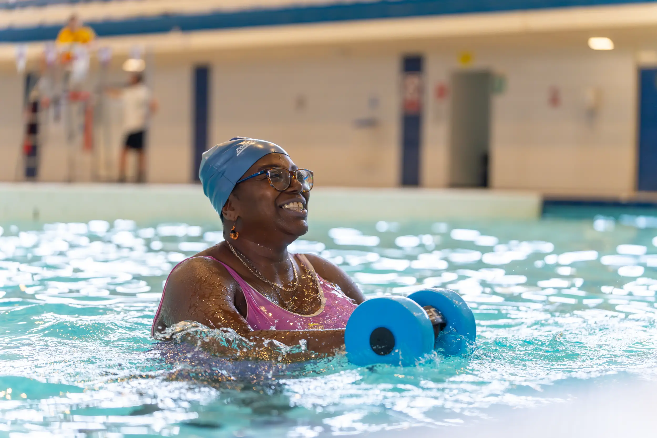 A smiling woman wearing a blue swim cap and glasses doing aqua aerobics in a swimming pool, holding blue foam dumbbells.