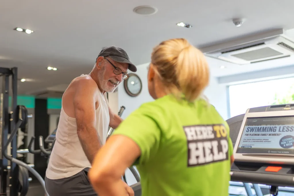 An elderly gentlemen, wearing a grey cap, having a gym assessment with a lady with blonde hair in a ponytail.