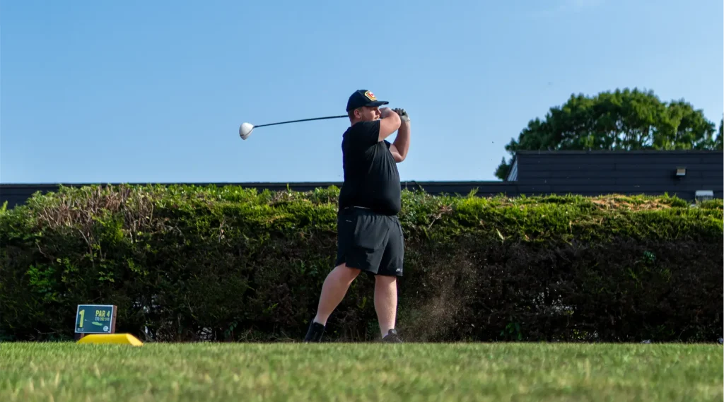 A young man, wearing a black cap, t-shirt and shorts, mind swing with a golf club.