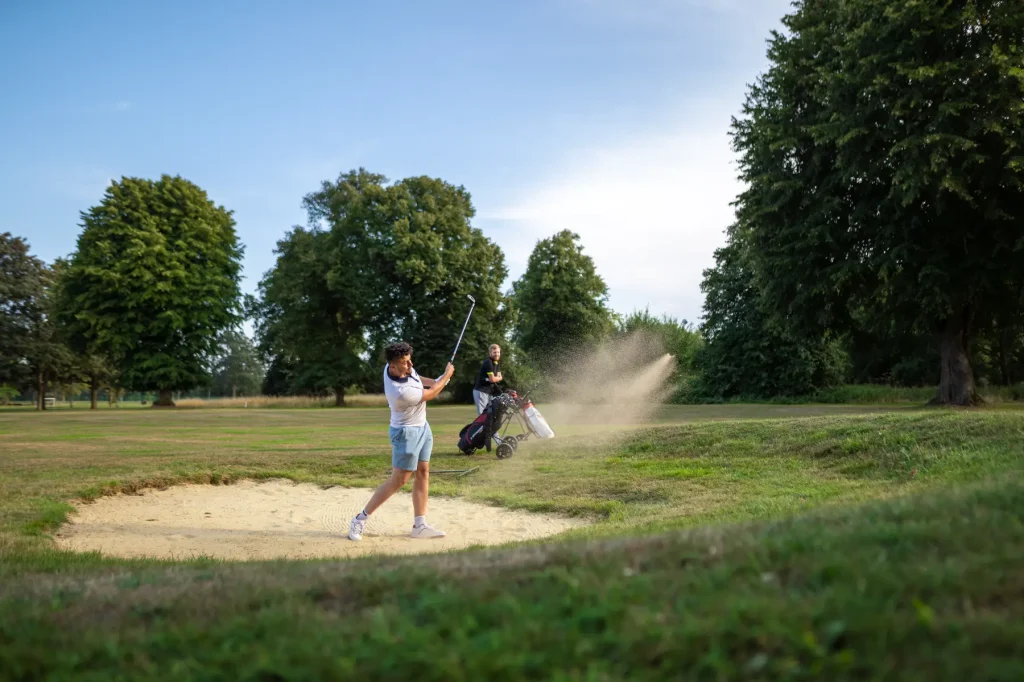A young man, wearing a white t-shirt and blue short, is mid swing taking his shot from the golf bunker. A man in black watches from the side lines with two golf caddys.