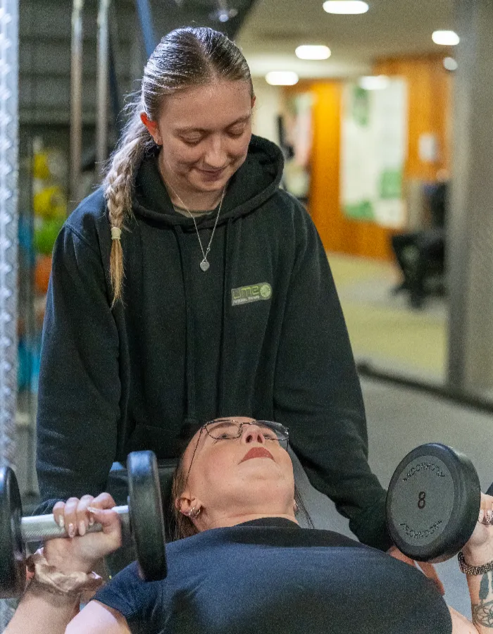 Lime personal trainer spotting a female client performing a dumbbell bench press exercise