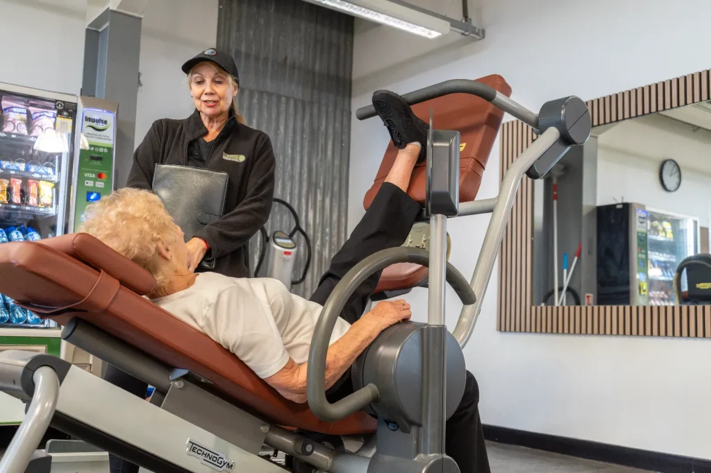 Lime personal trainer supporting an older lady using a Technogym leg press machine at Blackshots gym