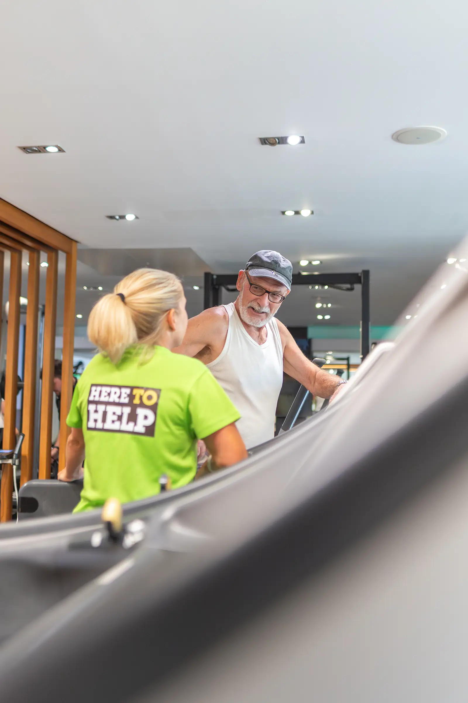 A female gym instructor, chatting to an elderly man on a treadmill in the gym.