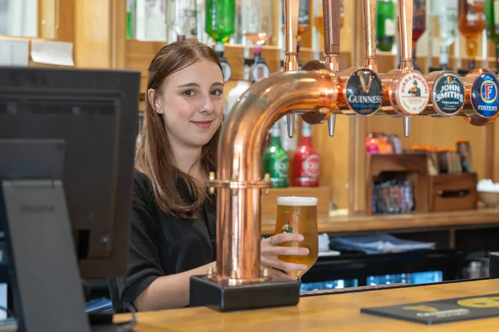 A young girl, smiling directly at the camera, pouring a pint behind the bar.