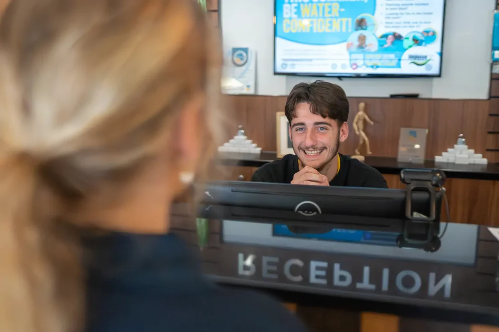 A young male receptionist, smiling at a lady with her hair in a pony tail.