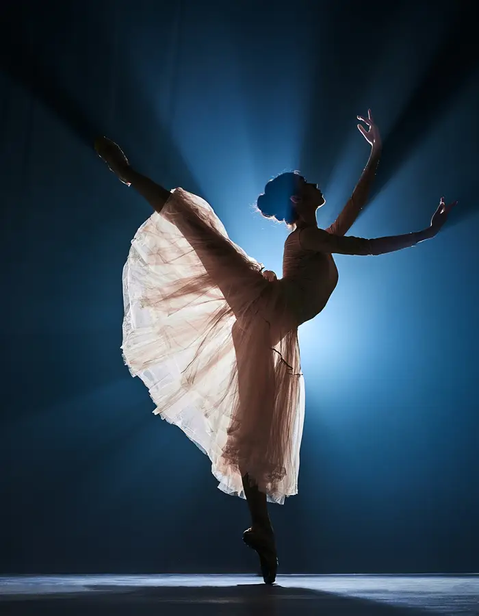 Silhouette of a ballerina in an arabesque pose on pointe, arms raised, backlit by dramatic blue stage lighting with a flowing skirt.