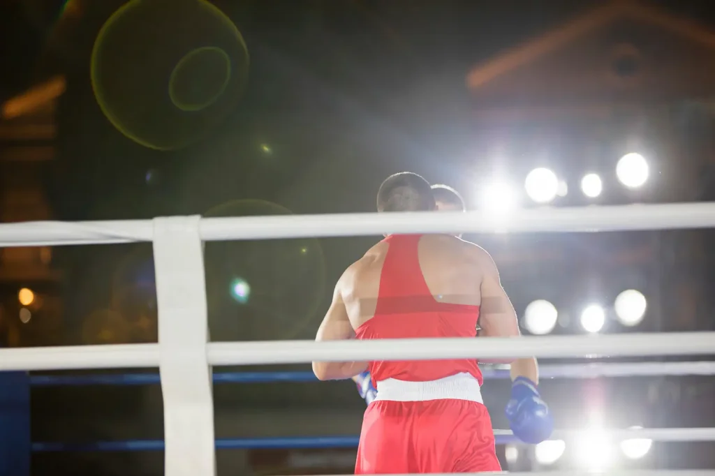 Close-up of an MMA fighter's gloved hand and flame-print shorts, standing in the cage with an opponent and crowd visible in the blurred background.