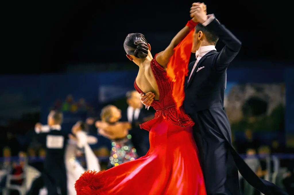 A ballroom dance couple mid-spin, the woman in a flowing red gown, with other competing couples visible blurred in the background.