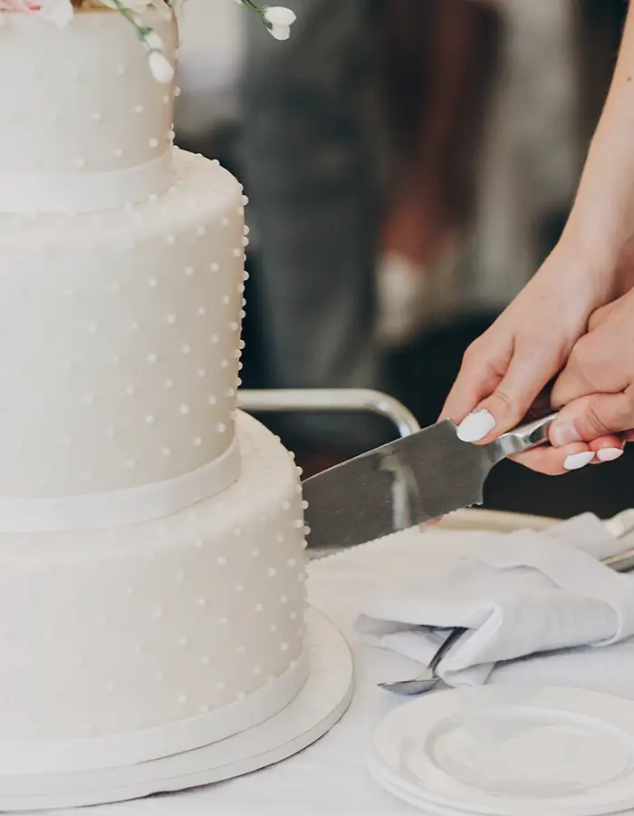 Close-up of hands cutting into a three-tier white wedding cake decorated with small beaded piping, with plates and a napkin-wrapped knife nearby.