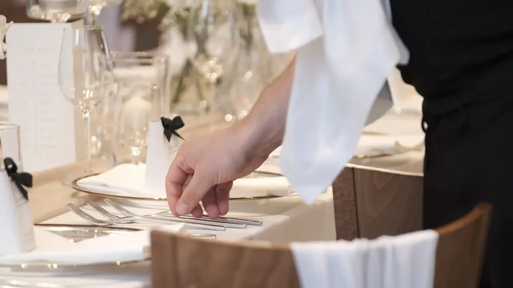 Close-up of a staff member's hand arranging cutlery and place settings at an elegantly laid wedding table with wine glasses, gold-rimmed plates, and a menu card.