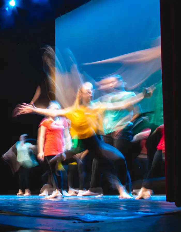 Long-exposure photo of a group of dancers on stage, their movements blurred into flowing streaks of colour under blue stage lighting.