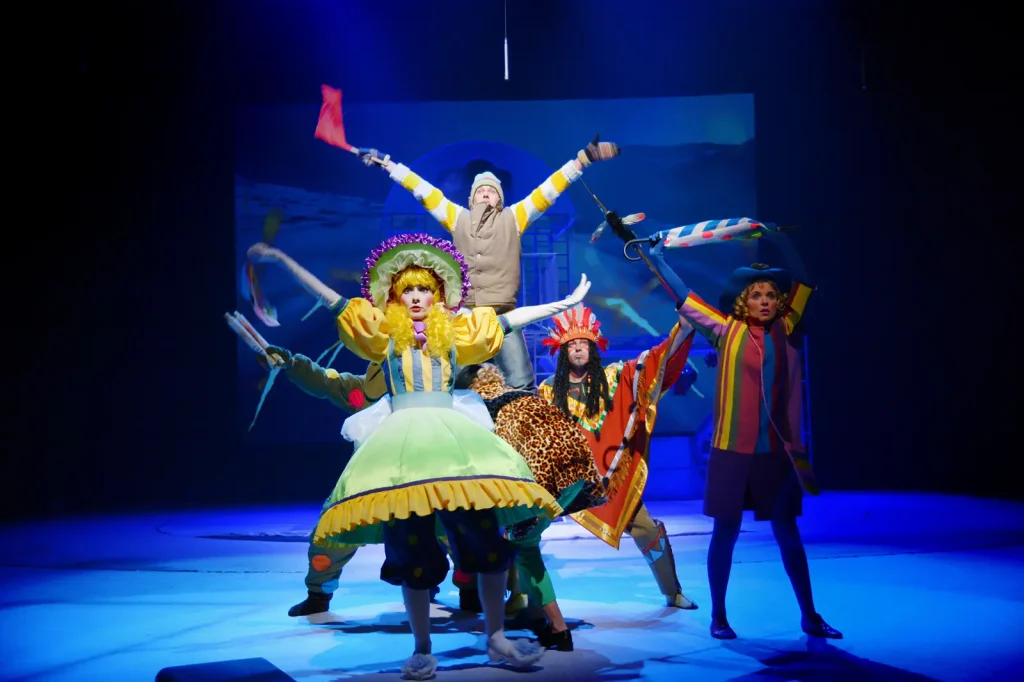 A group of costumed theatrical performers on stage in colorful, whimsical outfits — including a bonnet, a leopard-print cape, and a feathered headdress — striking playful poses under blue lighting.