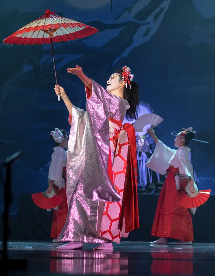 A performer in a pink kimono holding a red and white paper parasol, with two dancers in red and white kimonos and fans in the background, against a painted mountain backdrop.