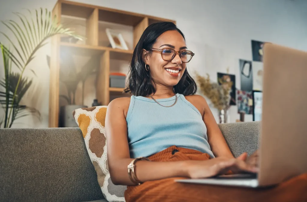A smiling woman with glasses sitting on a sofa at home, using a laptop.