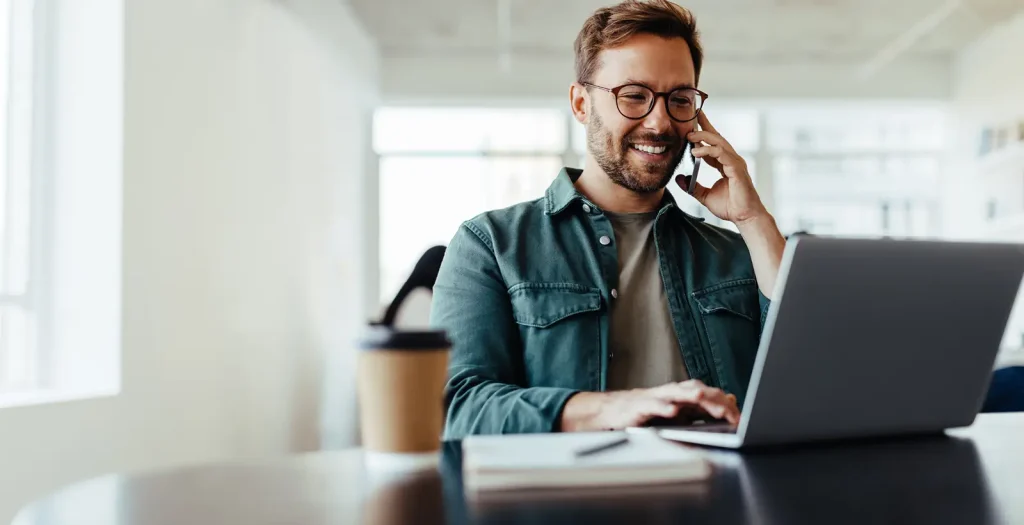 Man smiling on a phone call while working on a laptop in an office