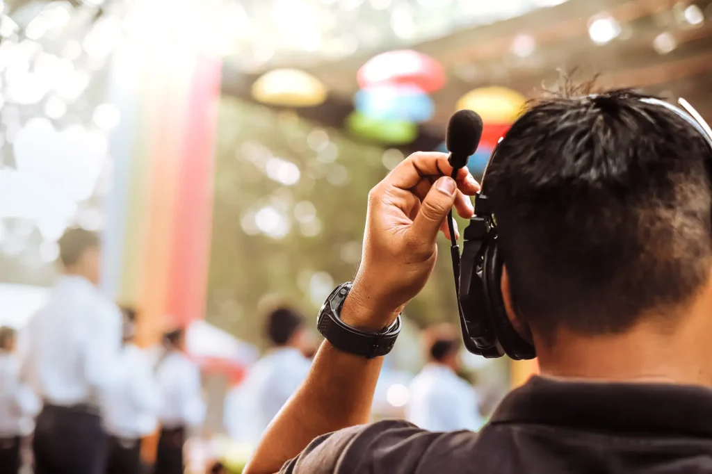Close-up from behind of a person adjusting a headset microphone at an outdoor event, with a blurred crowd and string lights in the background.