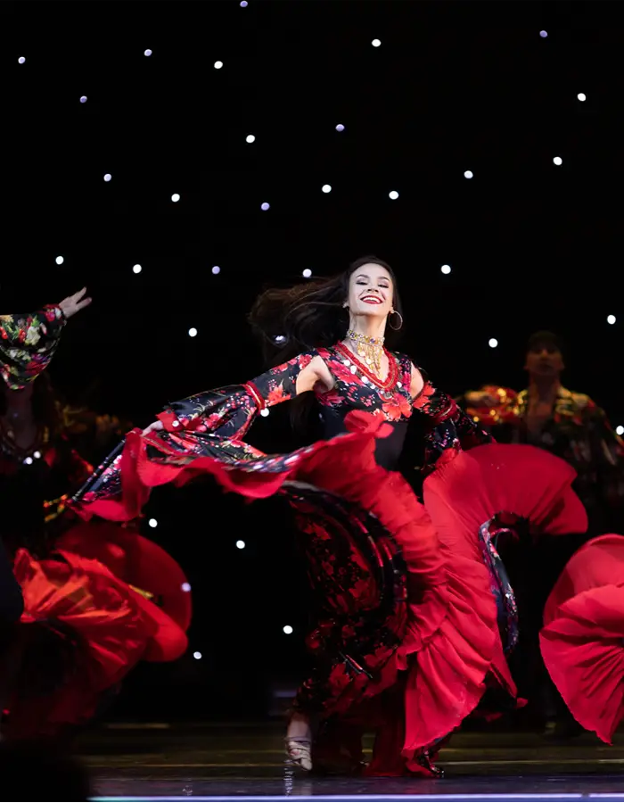 A smiling flamenco dancer in a red and black floral dress with a dramatic ruffled train, mid-spin on stage with other dancers in similar costumes behind her.