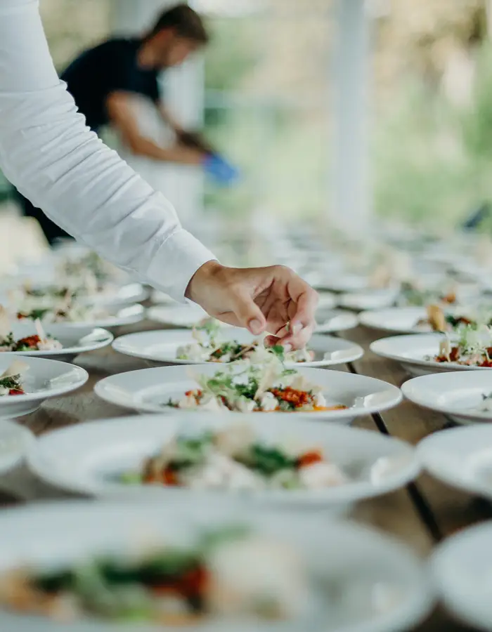 A caterer's hand garnishing rows of plated starters laid out on a long table, with another staff member working in the blurred background.