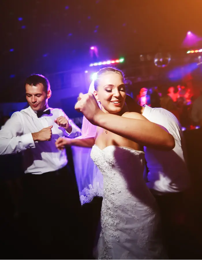 A bride in a strapless lace gown and veil dancing energetically at her reception, with a groomsman in a bow tie dancing behind her under colourful party lights.