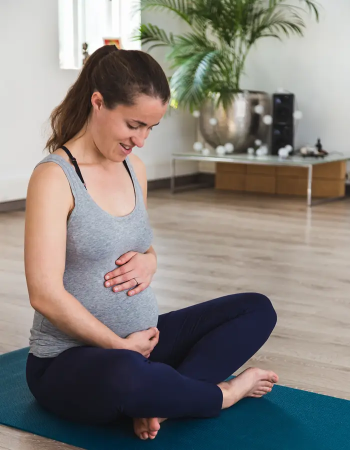 Pregnant lady in gym kit, sat crossed legged on a yoga mat, looking down cradling her bump.